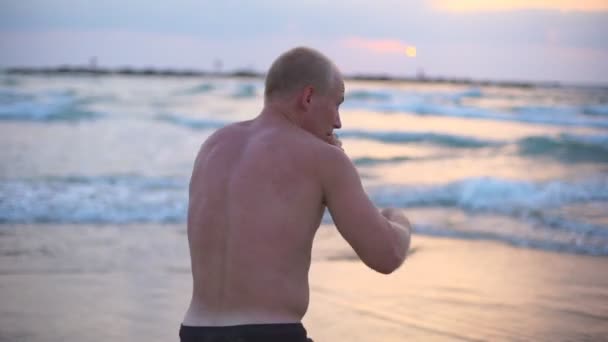 Junger muskulöser Mann beim Boxen am Strand des Meeres. Männliche Sportler üben sich allein in Selbstverteidigung in Küstennähe. Training von Kampfkünsten im Freien bei Sonnenuntergang. Zeitlupe Nahaufnahme — Stockvideo