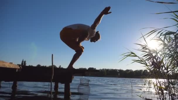 年轻男子站在湖边的木制防波堤边做瑜伽姿势。那个体格健壮的家伙在室外锻炼.背景为夏季风景。健康积极的生活方式的概念。近景侧景 — 图库视频影像