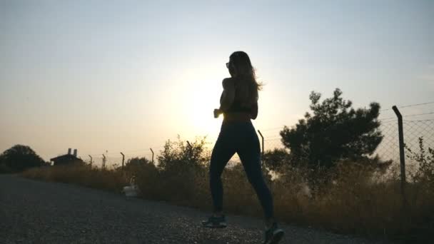 Mengikuti gadis sporty joging di jalan negara saat matahari terbit. Wanita muda berjalan di luar ruangan di pagi hari. Gaya hidup aktif sehat. Gerakan lambat — Stok Video