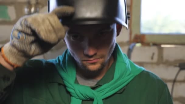 Sluiten van vermoeide jonge reparateur opent beschermend masker na afloop van zijn werk. Lasser in werkkleding op zoek naar camera in zijn garage. Portret van monteur met baard aan het werk in de werkplaats. Dolly schot — Stockvideo