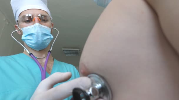 Medic met stethoscoop luistert longen van patiënt kijkt voor COVID-19 symptomen. Dokter met een onderzoekende man met stethoscoop aan coronavirus longontsteking tekenen. Artsen controleren van de borst van een man in het ziekenhuis — Stockvideo