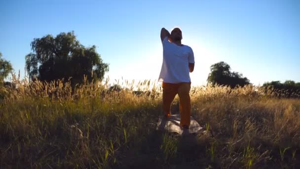 Mladý muž cvičí jógu na rohoži na louce za slunečného dne. Sportovní trénink na hřišti. Krásná krajina na pozadí. Koncept zdravého aktivního životního stylu. Nízký úhel pohledu Zavřít — Stock video