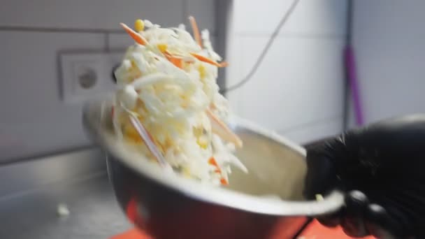 Mano masculina del chef en guante arrojando ingredientes y mezclándolos en un tazón de hierro. Brazo de cocinero revolviendo repollo chino, maíz, huevos y palitos de cangrejo para ensalada. Concepto de preparación de alimentos. Dolly disparó de cerca — Vídeo de stock