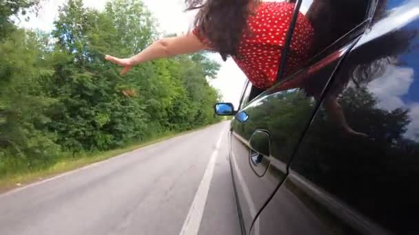 Brunett kvinna lutar sig ur att flytta bil och viftar med handen när hon rider genom landsvägen. Ung flicka sticker ut genom bilfönstret och hennes långa bruna hår blåser på vinden. Kvinnliga njuter av road trip — Stockvideo