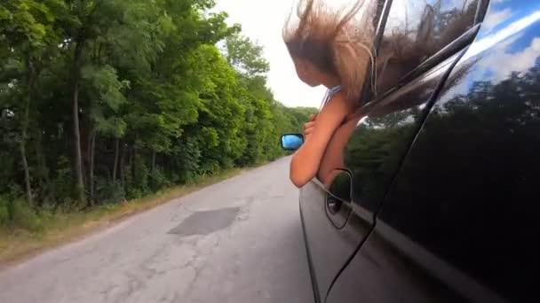 Little female child looks out of opening car window and enjoys trip riding through country road. Small girl sticks her head out of moving auto and her long blonde hair blows in wind. Rear view Slow mo — Stock Video