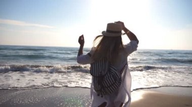 Sahilde koşan, bikinili ve sırt çantalı mutlu bir kız. Okyanus kıyısında koşan güzel bir kadın. Genç bayan hayattan zevk alıyor ve eğleniyor. Arka plan görünümünü kapat Yavaş çekim