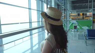 Sırt çantalı genç bir kadın havaalanı terminalinde duruyor ve pencereye bakıyor. Kız uçağını bekliyor. Turizm ve seyahat konsepti. Arka görünüm Yavaş çekim