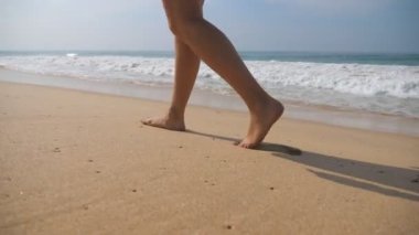 Kadın ayaklar arka planda okyanus dalgaları ile plajda altın kum üzerinde yürüyor. Genç kadının bacakları kuma basıyor. Deniz kıyısında yalınayak kız. Yaz tatili tatili. Yavaş hareket yakın çekim