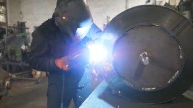 Atölyede koruyucu maske kaynakçısı. İki çelik detay arasında ortak çalışma yapan bir işçi. Adam metal parçalarını birbirine kaynaştırıyor. Metalle el emeği. Dolly vurdu.