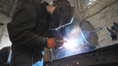 Metal parçalarını kaynaştıran işçiler. Kaynakçı iki çelik detay arasında ortaklık yapıyor. Metal kaynak yapan koruyucu maskeli bir adam. Endüstriyel üretim. Düşük açılı çekim
