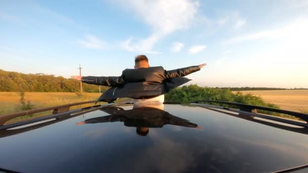 Young guy standing out of car sunroof with hands to sides and flying like plane while riding through country road. Man enjoying road trip and wind during journey. Travel or freedom concept. Back view — Stock Video