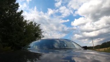 Siyah araba kırsaldan geçiyor. Kaputta yansımalar var. Gökyüzü, bulutlar ve ağaçlar. Otomobille otobanda gezmek manzaralı bir manzara. Kaputtan ön cama bak