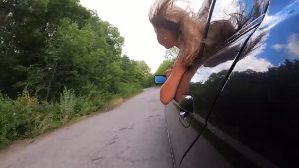 Petite fille regarde par la fenêtre d'ouverture de la voiture et aime voyager à travers la route de campagne. Petite fille sort sa tête de l'automobile en mouvement et ses longs cheveux blonds souffle dans le vent. Vue arrière Moteur lent — Video