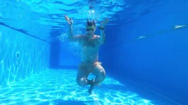 Individual transitions from walking to a seated position underwater in a clear blue pool, capturing the fluidity of movement and the tranquil ambiance of the aquatic scene