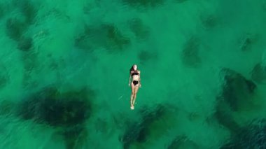 Woman in black swimwear is floating effortlessly on clear turquoise water, highlighting the serene atmosphere and the gentle ripples of the ocean in a captivating aerial view