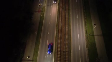 Aerial perspective captures vehicles traveling on a dimly lit road adjacent to railway tracks, illustrating motion and nighttime atmosphere, with camera following the scene fluidly
