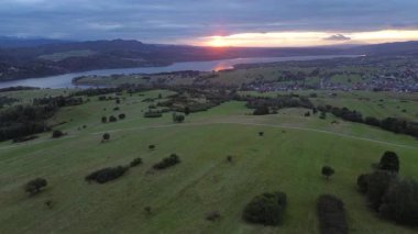 Aerial perspective captures tranquil landscape at sunset, featuring hills and a lake, as the camera smoothly zooms in, highlighting the vibrant colors and natural beauty