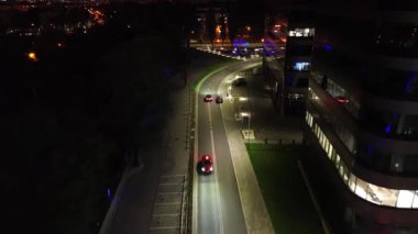 Gece modern bir ofis binası yakınında dolambaçlı bir yolda giden araba, çevre aydınlatması, dinamik hareketler ve şehir atmosferiyle aydınlatılmış, dramatik efekt için kamera tavaları ve eğilimleri olan bir araba.