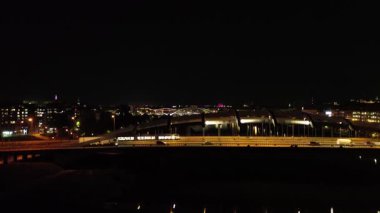 Gece şehir trafik manzarası aydınlanmış binaları ve akan araçları gösteriyor, dinamik bir kamera tavası canlı şehir yaşamını ve ufuk çizgisinde hareketliliği gösteriyor.