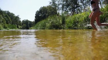 Genç bir kadın sığ nehir suyunda, canlı yeşilliklerle çevrili, yazın neşesini somutlaştıran ve doğanın huzur veren güzelliğini somutlaştıran