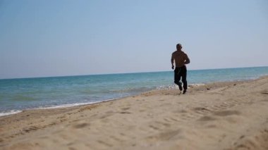 Adam kumlu bir sahil boyunca koşuyor, atletizm ve enerjiyi gösteriyor, dalgalar geliyor ve açık bir gökyüzü, açık hava egzersizi ve refahın ruhunu somutlaştırıyor.