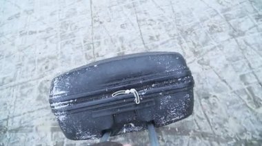 Karlı bir arazide manevra yapılan siyah bir valizin yakın çekimi, kar dokusu ve bavul özelliklerini vurgulayarak, kış manzaraları arasındaki yolculuğu gösteriyor