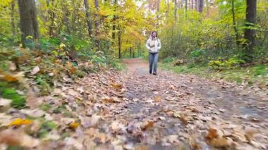 Genç bir kadın, canlı bir sonbahar ormanında yaprak kaplı yolda yürüyor, sahnenin doğal güzelliğini ve sükunetini gösteriyor, pürüzsüz bir kamera hareketiyle deneyimi geliştiriyor.
