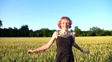 Pembe saçlı neşeli bayan hippi yeşil arpa tarlasında koşarken eğleniyor. Gün batımında buğday çayırında koşan dövmeli mutlu punk kadın. Neşeli hippi kız yaz gününün tadını çıkarıyor..