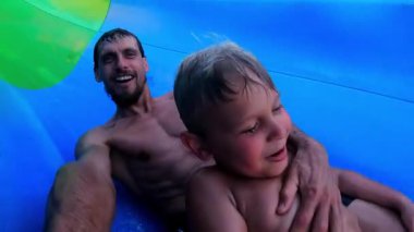 İnsan ve çocuk, renkli bir su kaydırağından kaymanın heyecanını yaşıyor. Neşeli ifadeler ve canlı renkler sergiliyor.