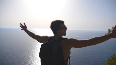 Sırt çantalı tanınmaz bir yürüyüşçü dağın kenarında duruyor ve muzaffer bir şekilde el kaldırıyor. Erkek turist yolculuk boyunca aktif olarak dinleniyor özgürlüğün ve güzel deniz manzarasının tadını çıkarıyor. Yavaş çekim Arka plan.