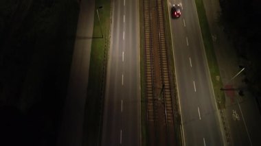 Demiryolu raylarının yanında karanlık bir yol boyunca giden bir arabanın hava görüntüsü, gece hareketlerini ve gece atmosferini gösteriyor, yavaş yavaş yakınlaştırarak çevreyi ortaya çıkarıyor.