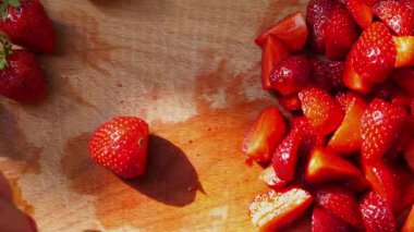 El, ahşap kesme tahtasındaki taze çilekleri ustalıkla dilimliyor. Canlı kırmızı meyve parçaları etrafa saçılıyor. Yemek hazırlama ve tazeliğin neşesini vurguluyor.