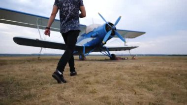 Pilot çimenli pistte klasik mavi çift kanatlı uçağa doğru yürüyor, heyecan verici bir uçuş deneyimi için hazırlanmanın heyecanını ve beklentisini yakalıyor.