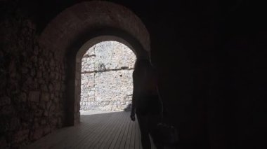 Güneş şapkalı bir kadın, gölgeden ışığa geçiş yapan antik bir taş kemer yolunda zarifçe ilerler. Keşif ve keşfin güzel bir yolculuğunu betimler.