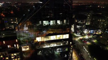 Akşam saatlerinde modern cam ofis binasının hava manzarası, aydınlanmış iç mekanlar ve çevre kentsel manzaralar, dramatik efektler için pürüzsüz bir kamera yakınlaşması.