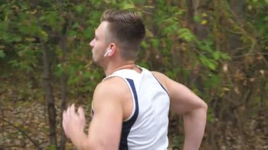 Genç sportif adam erken sonbaharda orman yakınında iz boyunca çalışan. Kablosuz kulaklıklar ile yakışıklı sporcu açık koşu. Atletik adam doğada egzersiz. Sağlıklı aktif yaşam tarzı. Yan görünüm.