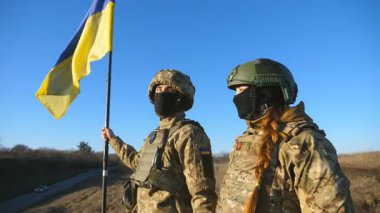 Ukrayna ordusunun genç askerleri ulusal bayrağıyla tepenin zirvesinde dikiliyorlar. Kamuflaj üniformalı kadın ve adam mavi gökyüzüne karşı Ukrayna bayrağını kaldırmış uzaklığa bakıyor. Kapat..