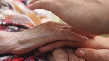 Yaşlı büyükannesinin yaşlı ellerini okşayan genç bir erkeğin yan görüntüsü. Aile dışarıda vakit geçiriyor. Şefkatli ve sevgi dolu bir kavram. Yavaşça kapat..