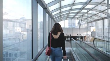 Genç bir kadın bavuluyla terminal koridorundan geçiyor. Yürüyen merdivende yürüyen ve tekerlekli bavulu yuvarlayan tanınmamış bir kız. Bayan seyahate ya da iş gezisine çıkar. Arka plan Yavaş çekim Kapat.