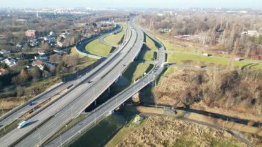 Yoğun bir otoyol kavşağının hava görüntüsü hareket halindeki gösteriş araçları, yemyeşil manzaralar ve arka planda kentsel alanlar, kamera olay yerinde sorunsuz bir şekilde yankılanıyor