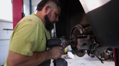 Yeşil tişörtlü yetenekli otomotiv tamircisi bir aracın fren tamiri ile meşgul, iyi donanımlı bir garaj ortamında araçlarla detaylı çalışmalar sergiliyor, işçiliği ve hassasiyeti vurguluyor.