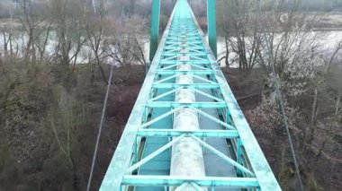 Metal iskeleti olan, uzun ve dar bir köprünün manzarası yükseldi ve sakin bir nehrin üzerine uzanan destek kirişleri etrafındaki ağaçları ve manzarayı sürekli olarak yansıtıyor.