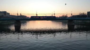 Gün batımında nehrin üzerindeki köprüden geçen tramvayın hava görüntüsü arabaların yanından geçişi, dinamik hareketler ve akşam gökyüzünün canlı renkleri suya yansıyor.