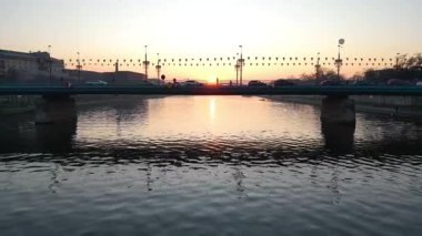 Gün batımında köprüden geçen arabaların hava görüntüsü canlı şehir yaşamını gözler önüne seriyor. Su ve aydınlık sokak lambaları kentsel manzarayı güçlendiriyor.