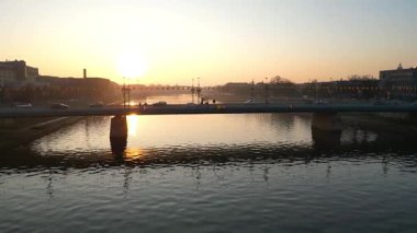 Gün batımında köprüden geçen arabaların hava görüntüsü canlı şehir yaşamını gözler önüne seriyor. Su ve aydınlık sokak lambaları kentsel manzarayı güçlendiriyor.