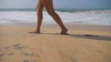 Kadın ayaklar arka planda okyanus dalgaları ile plajda altın kum üzerinde yürüyor. Genç kadının bacakları kuma basıyor. Deniz kıyısında yalınayak kız. Yaz tatili tatili. Yavaş hareket yakın çekim.