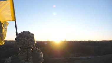Ukraynalı asker tepenin zirvesinde gün batımına karşı mavi-sarı bayrak sallıyor. Kamuflaj üniformalı erkek asker Ukrayna bayrağını kaldırıyor. Rus saldırısına karşı zafer simgesi olarak..