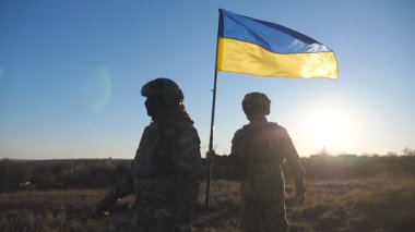 Askeri üniformalı genç kadın ve adam gün batımında çayırda mavi-sarı pankartıyla gidiyorlar. Ukrayna bayrağıyla yürüyen Ukrayna ordusunun kadın ve erkek askerleri. Rus işgaline direniş.
