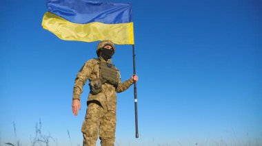 Mavi-sarı pankartıyla mavi gökyüzüne selam veren Ukraynalı erkek asker. Kırsalda Ukrayna bayrağı taşıyan kamuflaj üniformalı genç bir asker. Rus istilasına direniş. Düşük görünüm.