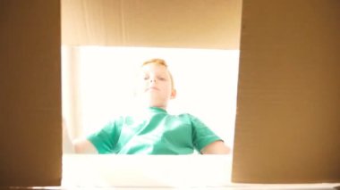 Yüzünde olumlu duygular ve duygular olan bir paket kutusuna bakan kızıl saçlı küçük bir çocuk. Genç bir çocuk karton kutuyu açıyor ve içinden hediyesini çıkarıyor. Küçük çocuk neşe ve şaşkınlık gösteriyor. Yavaş çekim.